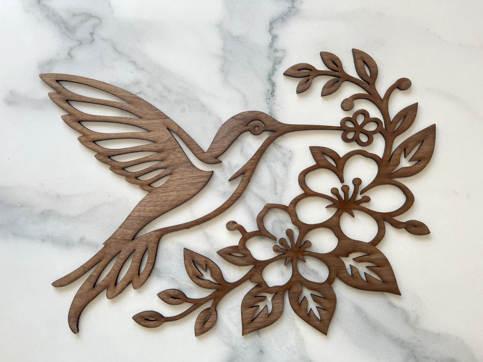 Décoration en bois de noyer noir représentant un colibri en vol entouré de fleurs, de feuilles et de petites branches finement découpées au laser.