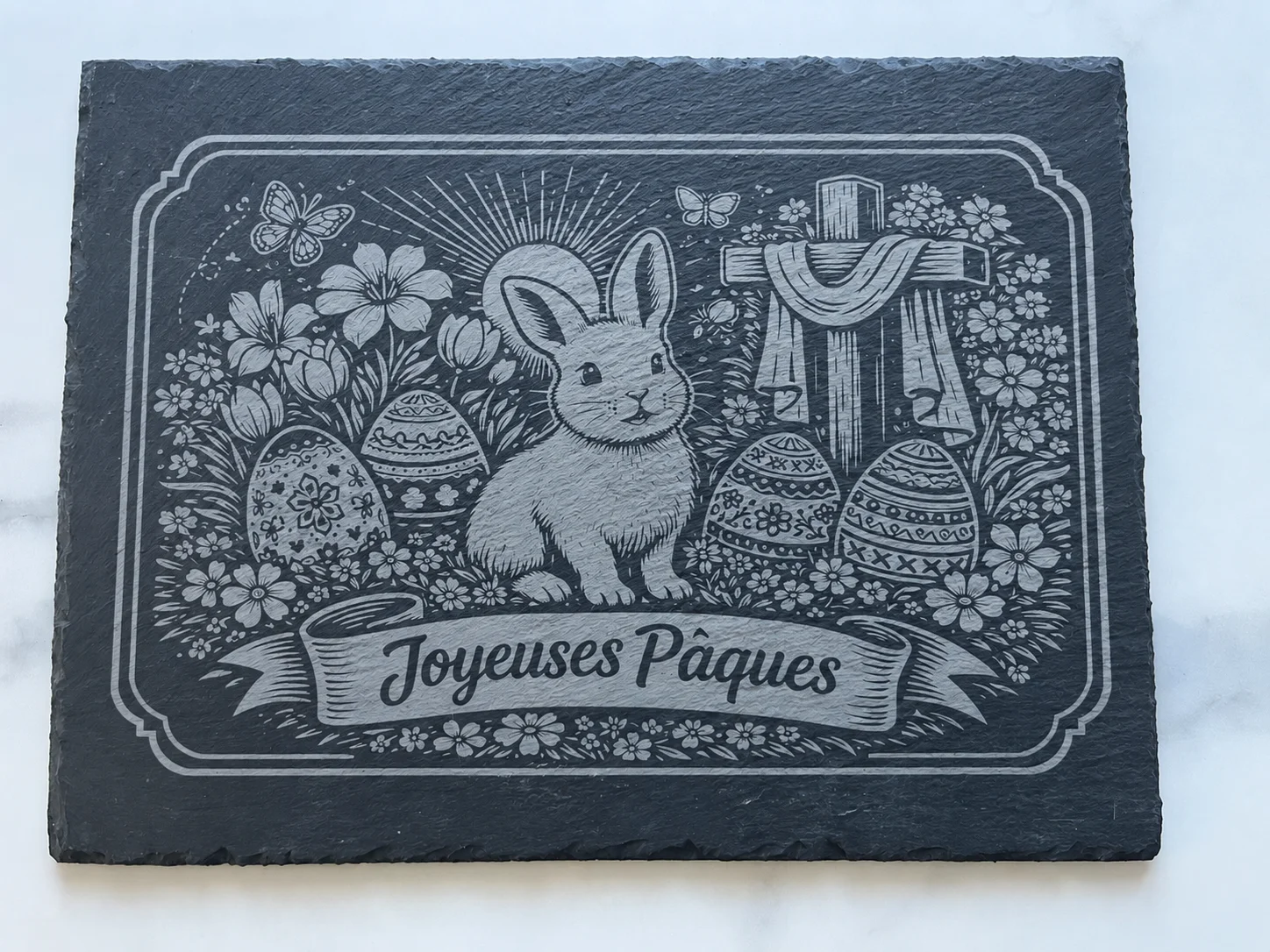Ardoise noire gravée d’un lapin de Pâques entouré d’œufs, de fleurs, de papillons et d’une croix, avec l’inscription « Joyeuses Pâques ».